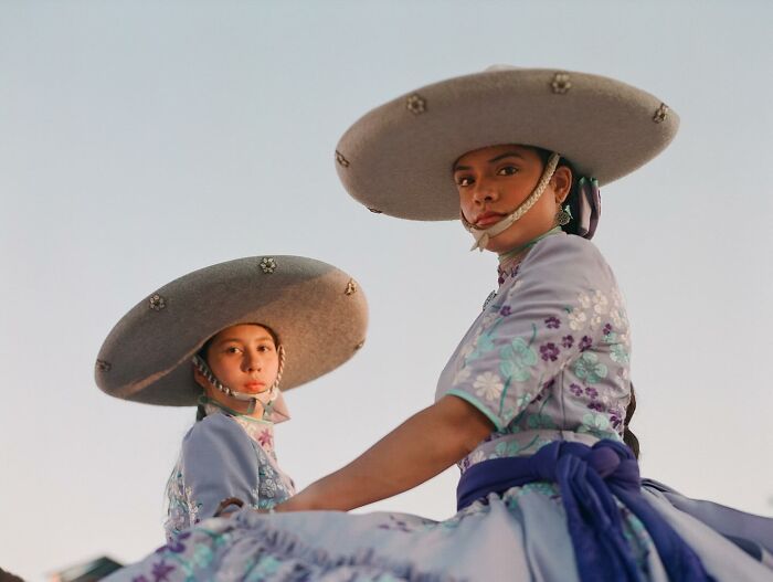 Two all-female rodeo riders wearing traditional dresses and large sombrero hats, captured beautifully outdoors.