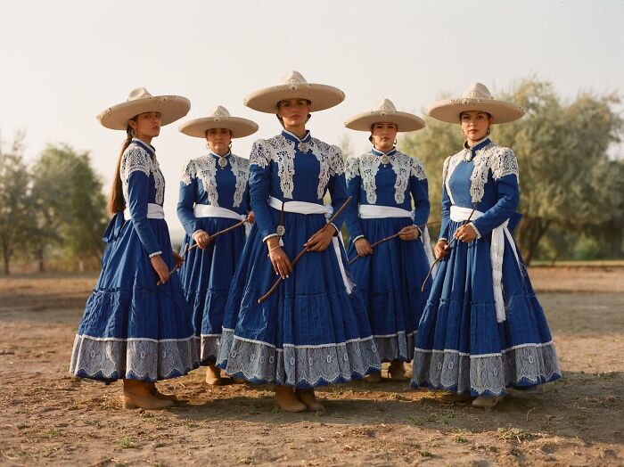 All-female rodeo riders in traditional blue dresses and wide-brimmed hats posing outdoors at sunset.