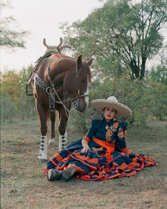 Female rodeo rider in traditional attire sitting on grass beside a saddled horse in a natural outdoor setting.