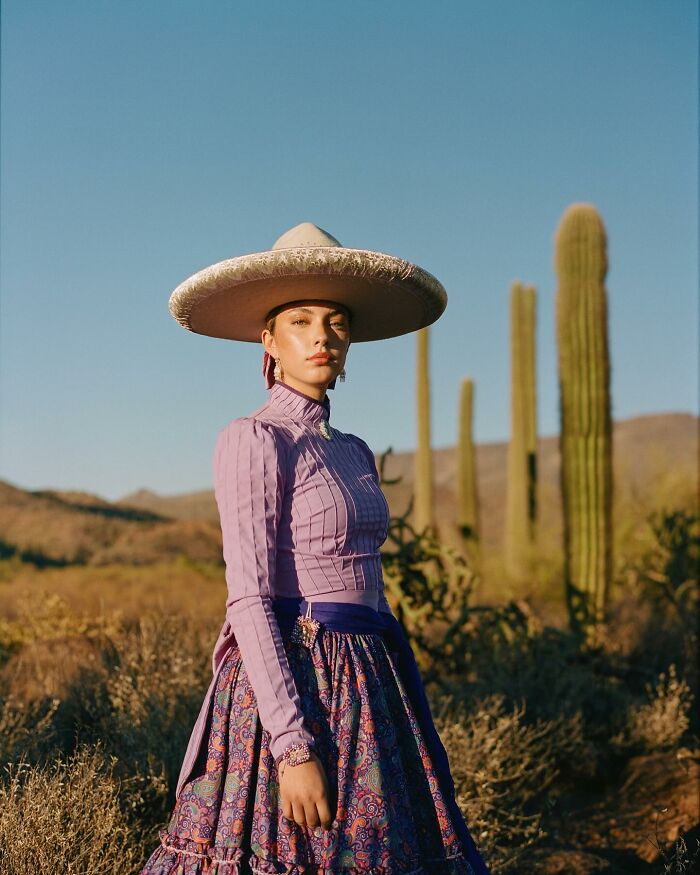 Female rodeo rider wearing traditional attire and a wide-brimmed hat standing in a desert landscape with cacti.