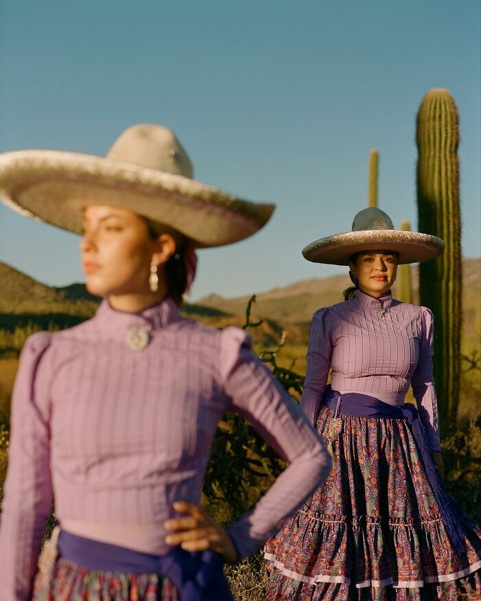 Two all-female rodeo riders wearing traditional dresses and wide-brimmed hats in a desert landscape with cacti.