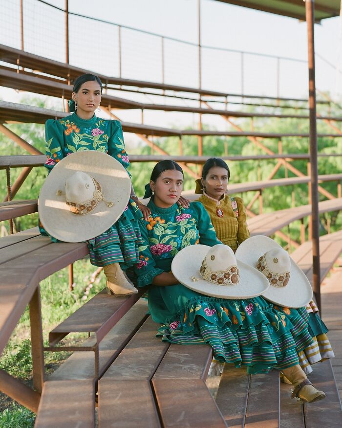 Three all-female rodeo riders in traditional outfits holding wide-brimmed hats, sitting on wooden bleachers outdoors.