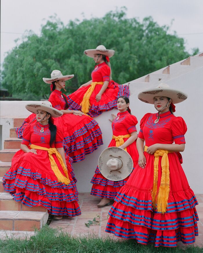 All-female rodeo riders posing in vibrant traditional red dresses and wide-brimmed hats on outdoor stairs.