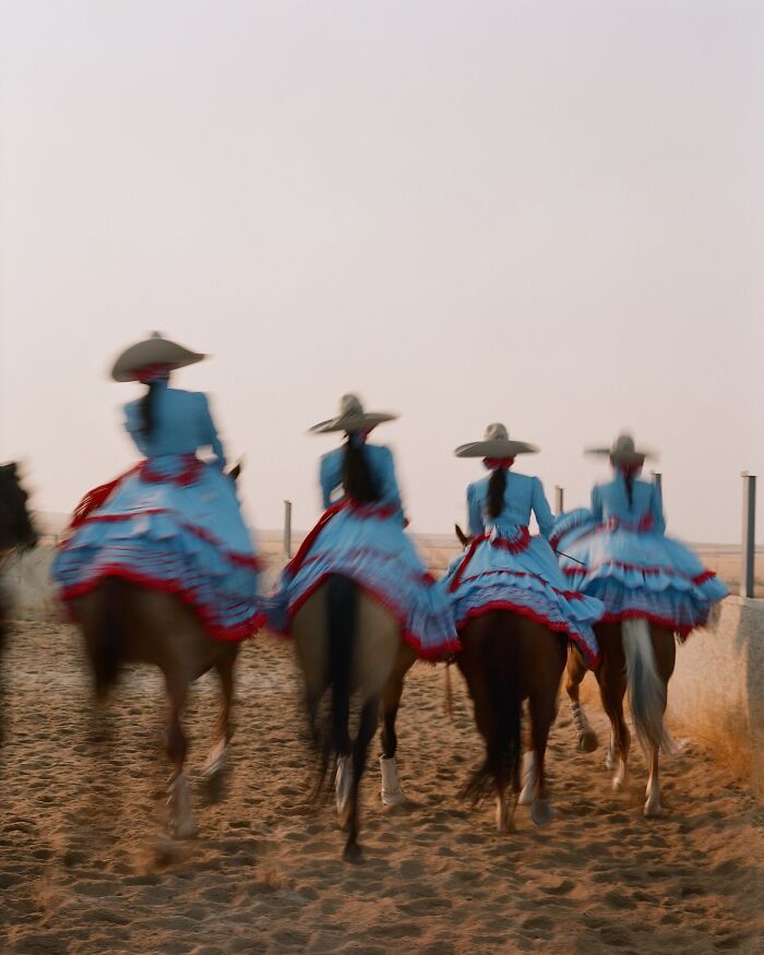 All-female rodeo riders in traditional dresses and hats riding horses in a sandy arena at sunset.