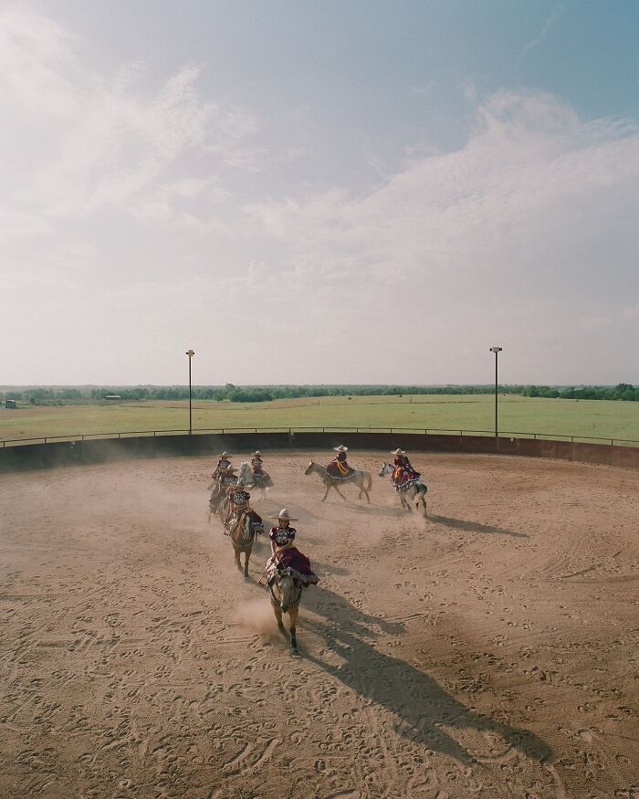 All-female rodeo riders on horseback riding in formation inside a dusty round arena on a sunny day.