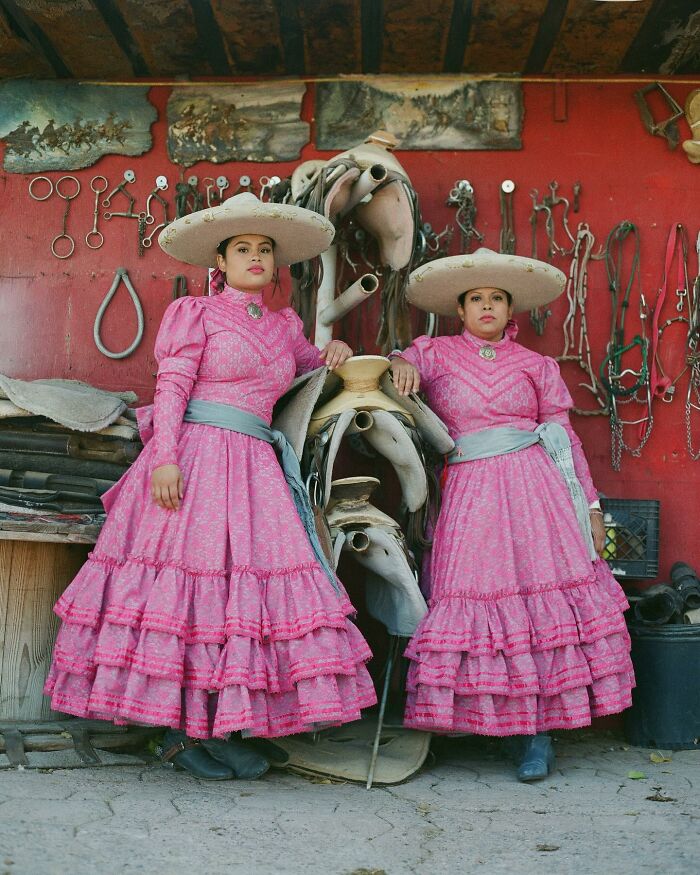 Two all-female rodeo riders in traditional pink dresses and wide-brimmed hats standing by saddles and tack.