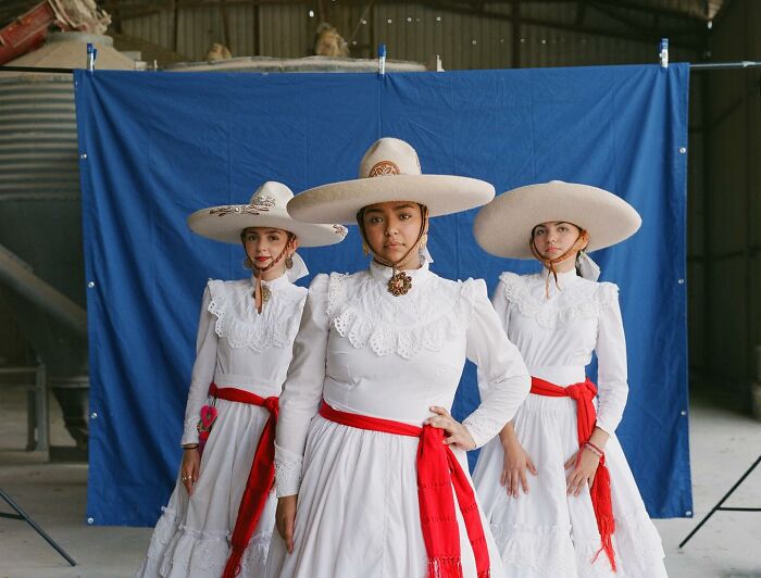 Three all-female rodeo riders in traditional white dresses and wide-brimmed hats posing against a blue backdrop.