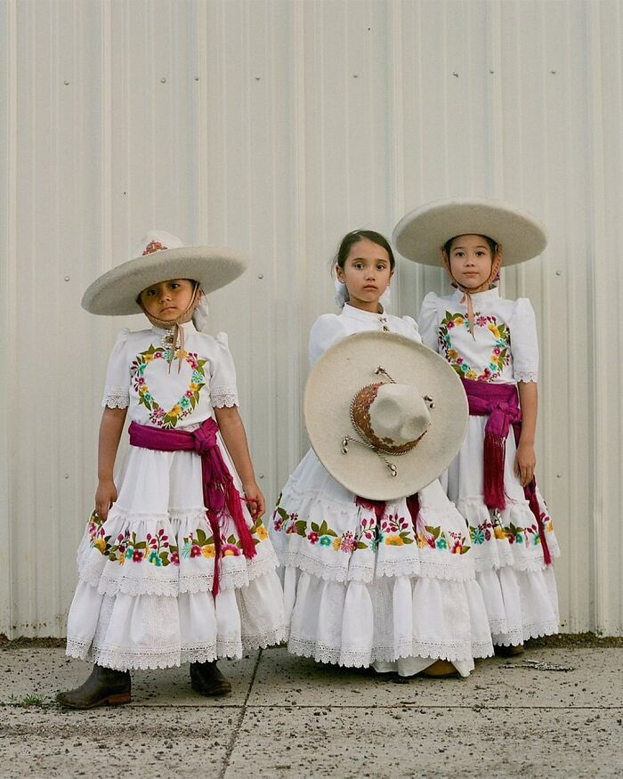 Three young all-female rodeo riders in traditional embroidered white dresses and large sombrero hats posing outdoors.