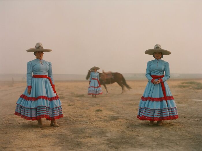 All-female rodeo riders in traditional blue and red dresses stand in a dusty arena with a horse and rider in the background.