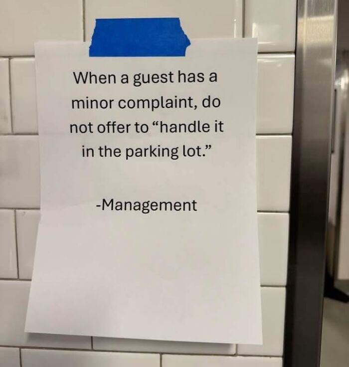 Sign on white tile wall with humorous hospitality meme about handling guest complaints professionally, taped with blue tape.