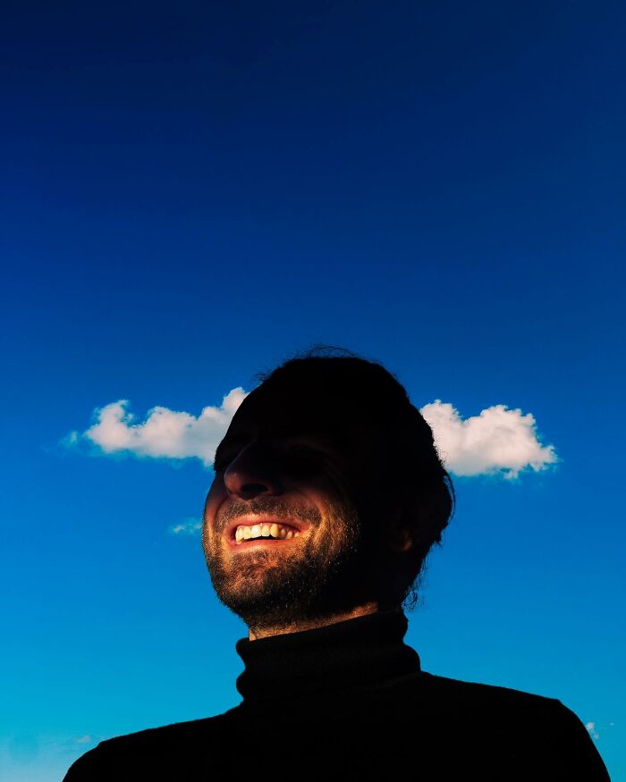 Man smiling against a vibrant blue sky with creative photo manipulations showing clouds behind his head in a surreal style.