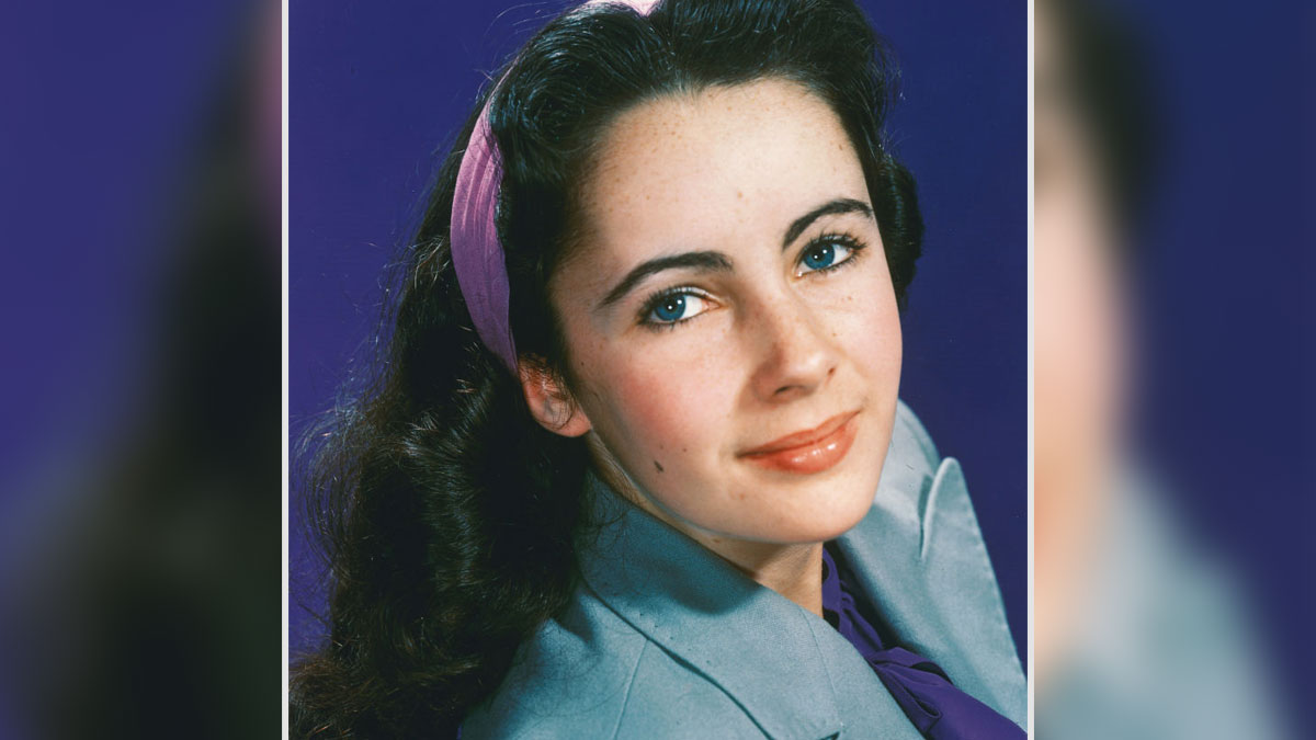 Young Elizabeth Taylor with striking blue eyes and dark hair wearing a gray jacket and purple headband in a studio portrait.