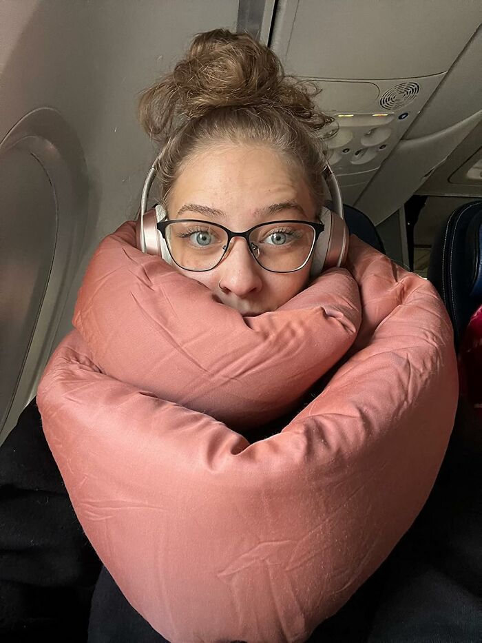 Woman on airplane wearing headphones and glasses, using a pink travel pillow for in-flight essentials comfort.