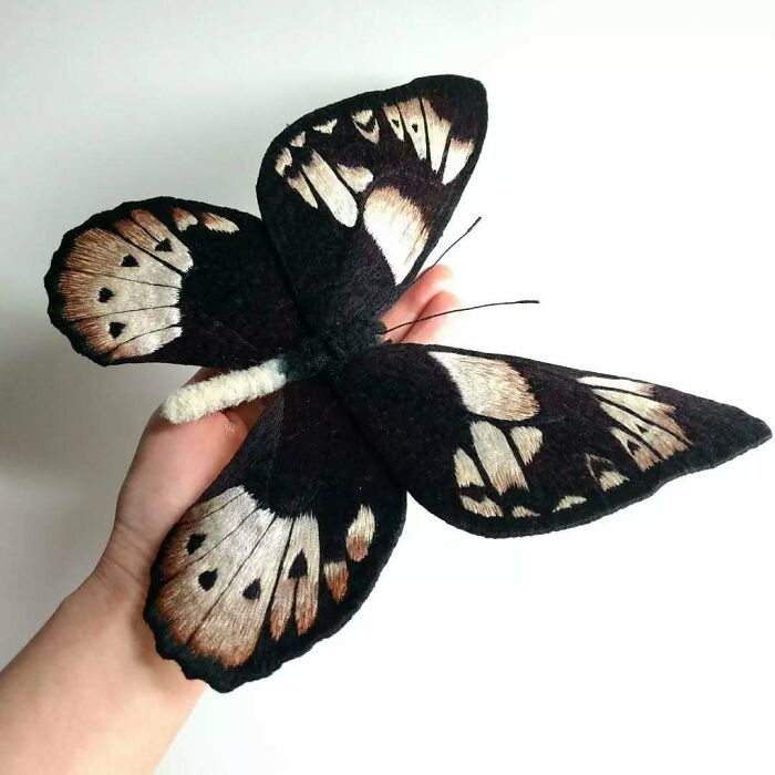 Hand holding a large, beautiful embroidered fiber moth with detailed black and beige wings by Yumi Okita.
