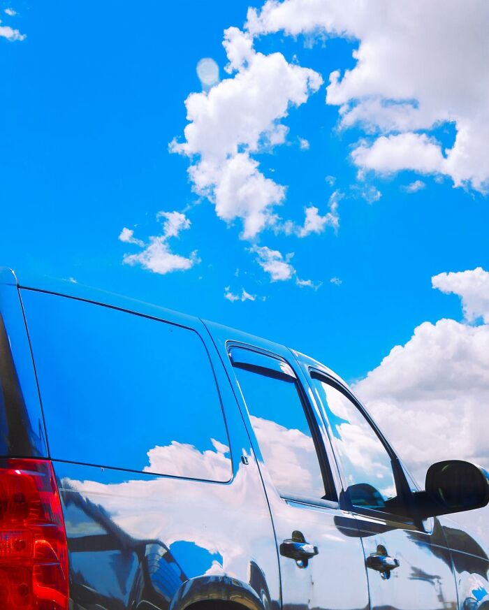Black car reflecting bright blue sky and white clouds, showcasing creative photo manipulations in a surreal reality style.