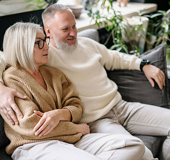 Mature couple sitting closely on a couch, showing concern and support amid wedding bully invitation conflict. Mature couple sitting closely on a couch, showing concern and support amid wedding bully invitation conflict.