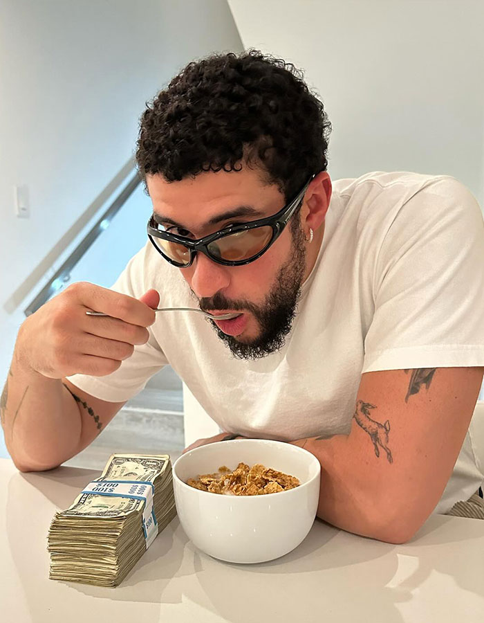 Bad Bunny wearing sunglasses, eating cereal at a table next to stacks of cash in a casual indoor setting. Bad Bunny wearing sunglasses, eating cereal at a table next to stacks of cash in a casual indoor setting.