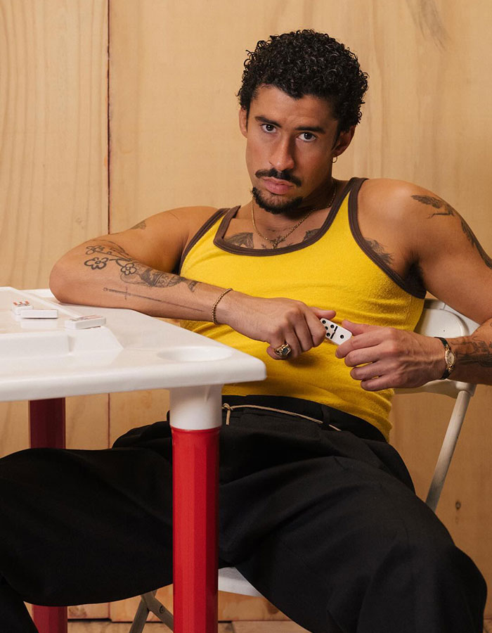 Man wearing a yellow tank top and black pants sitting at a table holding dominoes, embodying Bad Bunny style and vibe. Man wearing a yellow tank top and black pants sitting at a table holding dominoes, embodying Bad Bunny style and vibe.