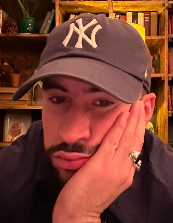 Close-up selfie of Bad Bunny wearing a New York Yankees cap, resting his face on his hand indoors with bookshelves behind. Close-up selfie of Bad Bunny wearing a New York Yankees cap, resting his face on his hand indoors with bookshelves behind.