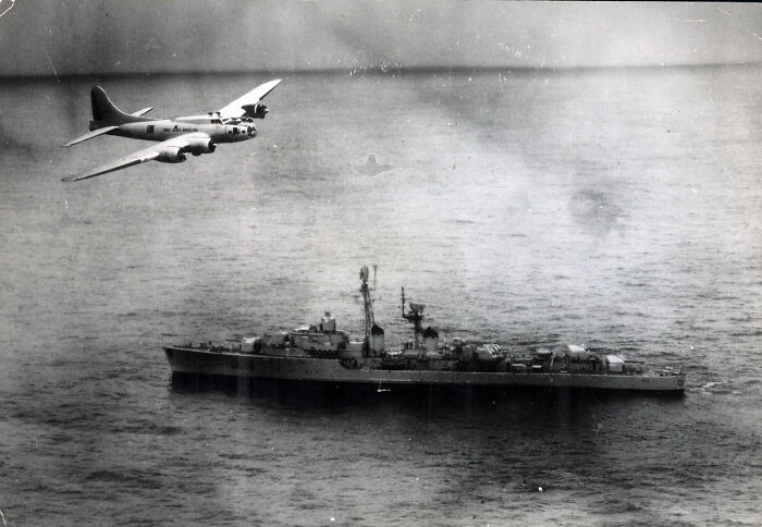 Vintage black and white photo of a military airplane flying over a battleship on the ocean, depicting bizarre historical events.
