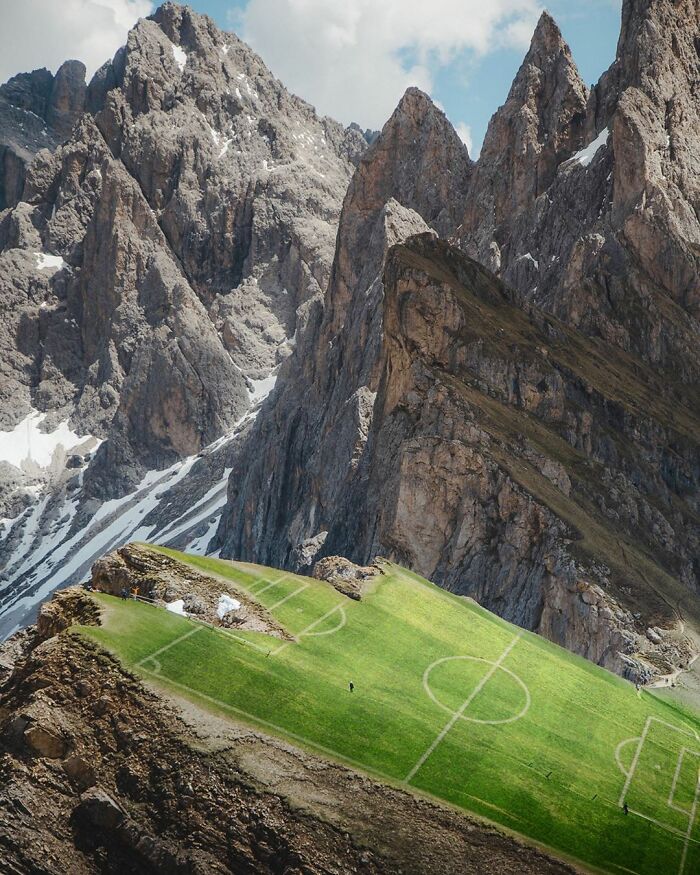 Photo manipulation showing a soccer field creatively placed on a grassy mountain slope amidst rocky peaks.
