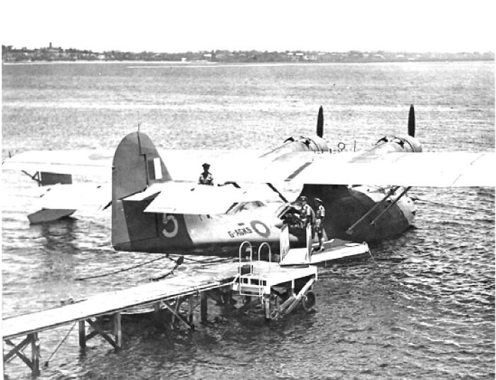 Bizarre historical events showing a vintage seaplane docked with crew on the wing by the water in black and white.