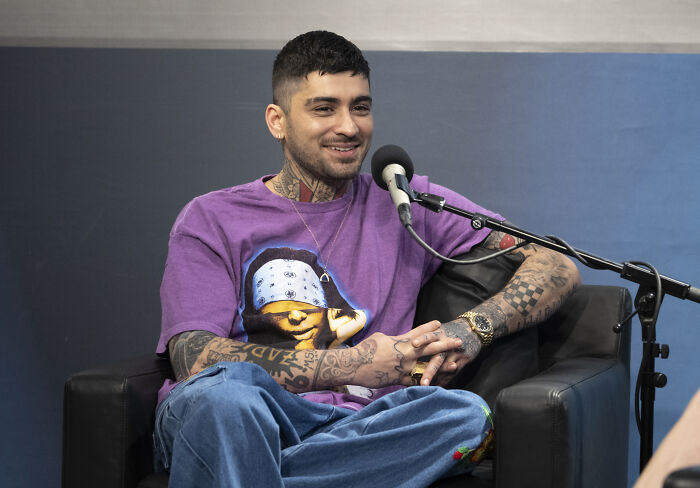 Former band member with tattoos and earrings, wearing a purple graphic tee, sitting and smiling during an interview session. Former band member with tattoos and earrings, wearing a purple graphic tee, sitting and smiling during an interview session.