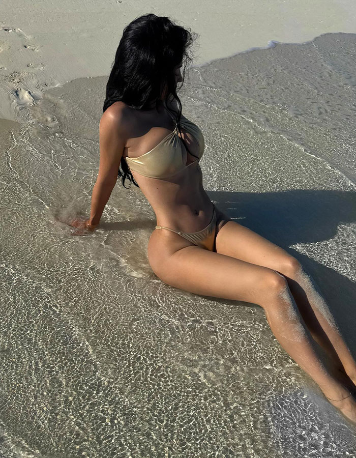 Woman in a revealing golden bikini sitting on wet sand, showcasing a round butt at the beach with clear shallow water around. Woman in a revealing golden bikini sitting on wet sand, showcasing a round butt at the beach with clear shallow water around.