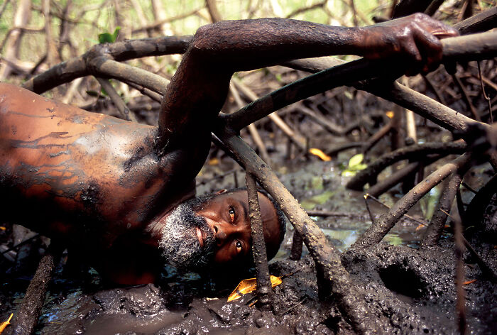 Man covered in mud lying among mangrove roots capturing unique nature moments for mangrove photography awards entries.
