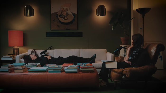 Controversial artist reclining on a couch during podcast interview in a dimly lit room with stacks of books nearby. Controversial artist reclining on a couch during podcast interview in a dimly lit room with stacks of books nearby.