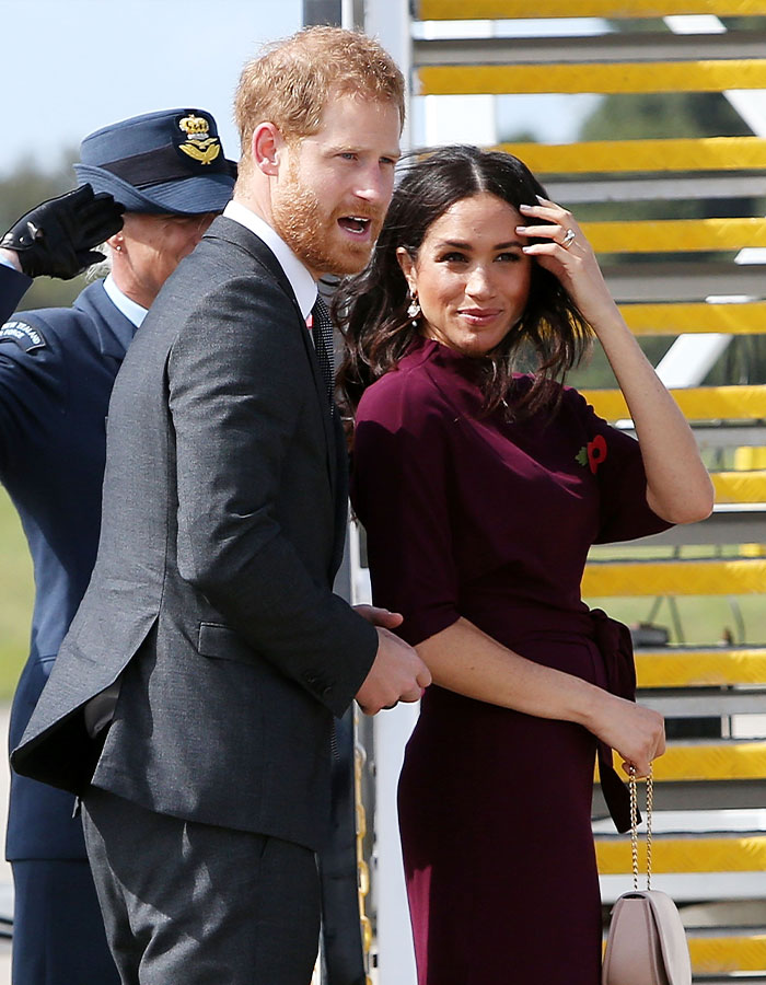 Meghan Markle smiling in a burgundy dress standing with Prince Harry near an aircraft staircase outdoors. Meghan Markle smiling in a burgundy dress standing with Prince Harry near an aircraft staircase outdoors.