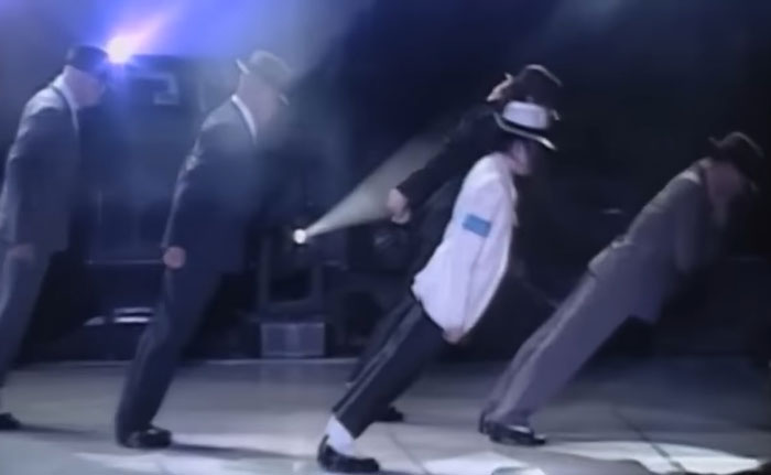 Michael Jackson performing his iconic lean move on stage with backup dancers in suits and fedoras. Michael Jackson performing his iconic lean move on stage with backup dancers in suits and fedoras.