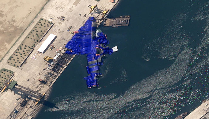 Aerial view of a 5,000-ton warship capsized near the dock, showing damage and debris in the water. Aerial view of a 5,000-ton warship capsized near the dock, showing damage and debris in the water.