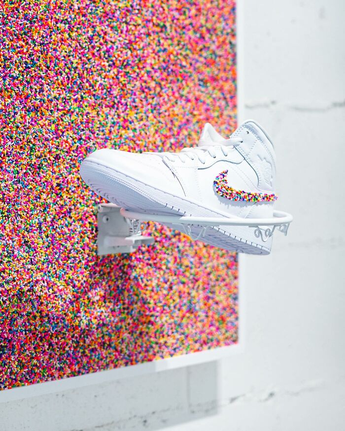 White sneaker with Nike logo made of colorful sprinkles mounted on a mini basketball hoop in sprinkle-covered art installation.