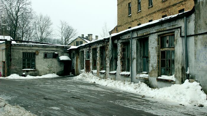 Abandoned urban alley covered with snow, showing weathered buildings, c*****d windows, and a gloomy winter atmosphere.