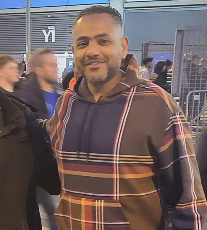 Man in a plaid hoodie smiling at a crowded outdoor event, representing father of four with few followers jailed. Man in a plaid hoodie smiling at a crowded outdoor event, representing father of four with few followers jailed.