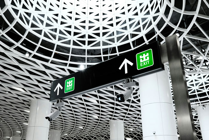 Airport exit sign under a modern ceiling structure, related to facial recognition issues with humiliated passenger makeup removal. Airport exit sign under a modern ceiling structure, related to facial recognition issues with humiliated passenger makeup removal.
