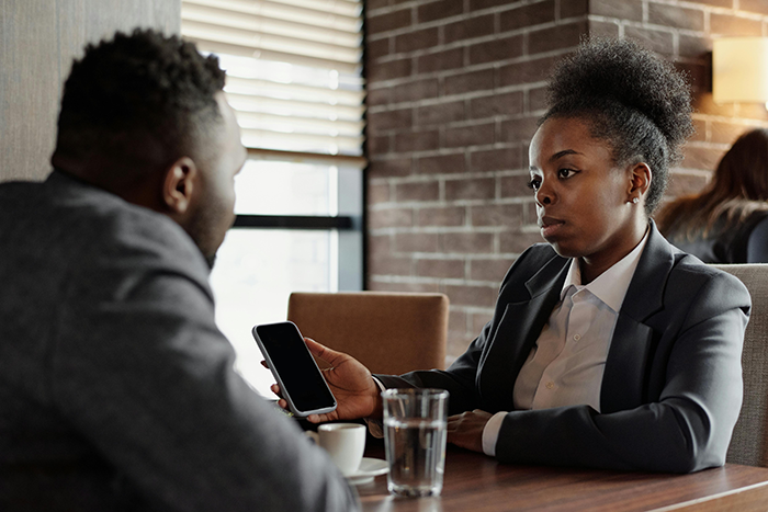 Woman in business attire shows phone to coworker in office, expressing shock as private dating profile is revealed. Woman in business attire shows phone to coworker in office, expressing shock as private dating profile is revealed.