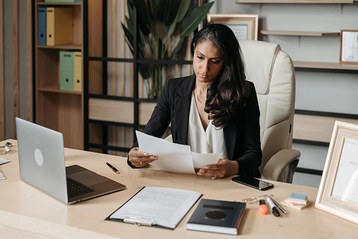 Woman at office desk looking distressed while reviewing documents, with laptop and phone nearby in a professional setting Woman at office desk looking distressed while reviewing documents, with laptop and phone nearby in a professional setting