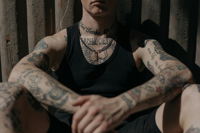 Person with multiple tattoos on arms and neck, sitting against a dark textured background in soft sunlight. Person with multiple tattoos on arms and neck, sitting against a dark textured background in soft sunlight.