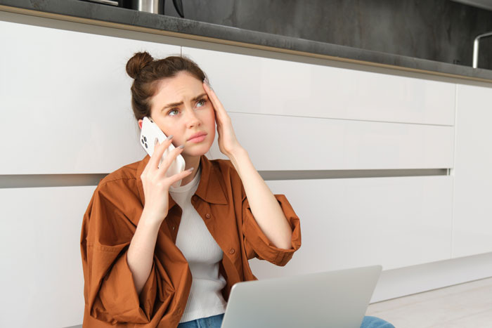 Young woman stressed on phone while sitting on floor near laptop, reflecting challenges of caring for kids alone. Young woman stressed on phone while sitting on floor near laptop, reflecting challenges of caring for kids alone.