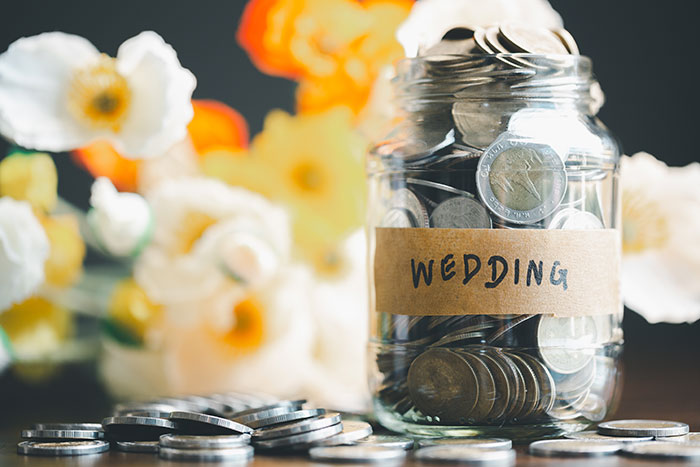Glass jar labeled wedding filled with coins surrounded by flowers, relating to paying for hen parties debates online. Glass jar labeled wedding filled with coins surrounded by flowers, relating to paying for hen parties debates online.