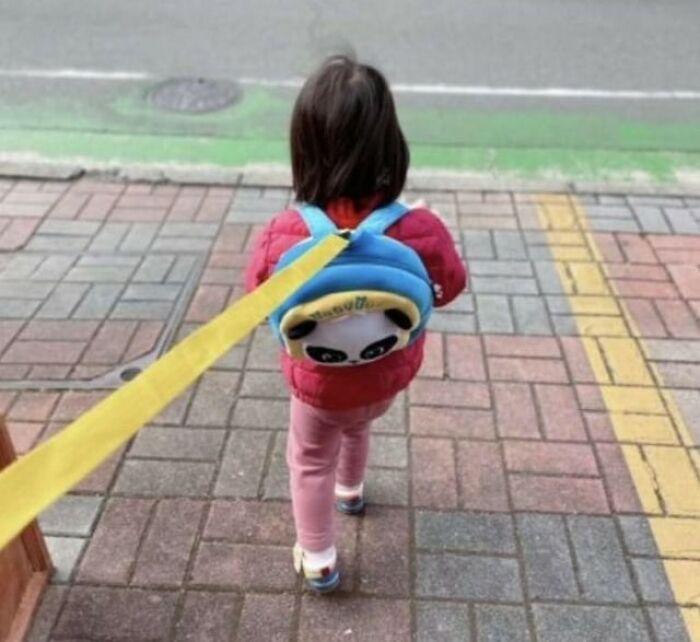Child wearing a panda backpack with a yellow leash walking on a paved sidewalk near a street curb.