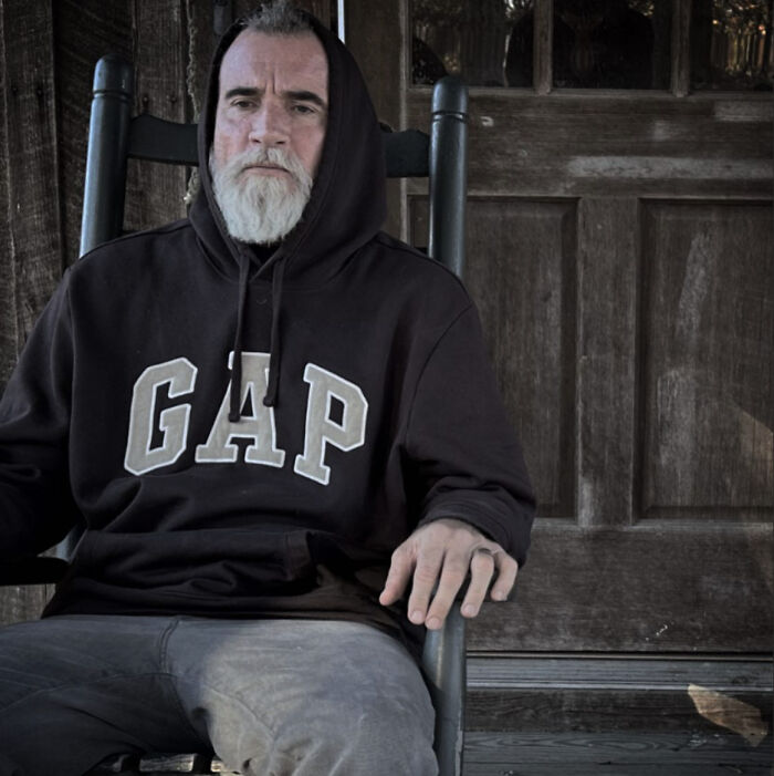 Dominic Purcell, Prison Break star, with a full white beard and hoodie, sitting on a chair in a rustic setting.