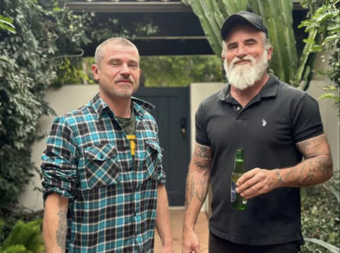 Dominic Purcell with a new beard and cap standing outdoors with a tattooed man in casual attire holding a green bottle.