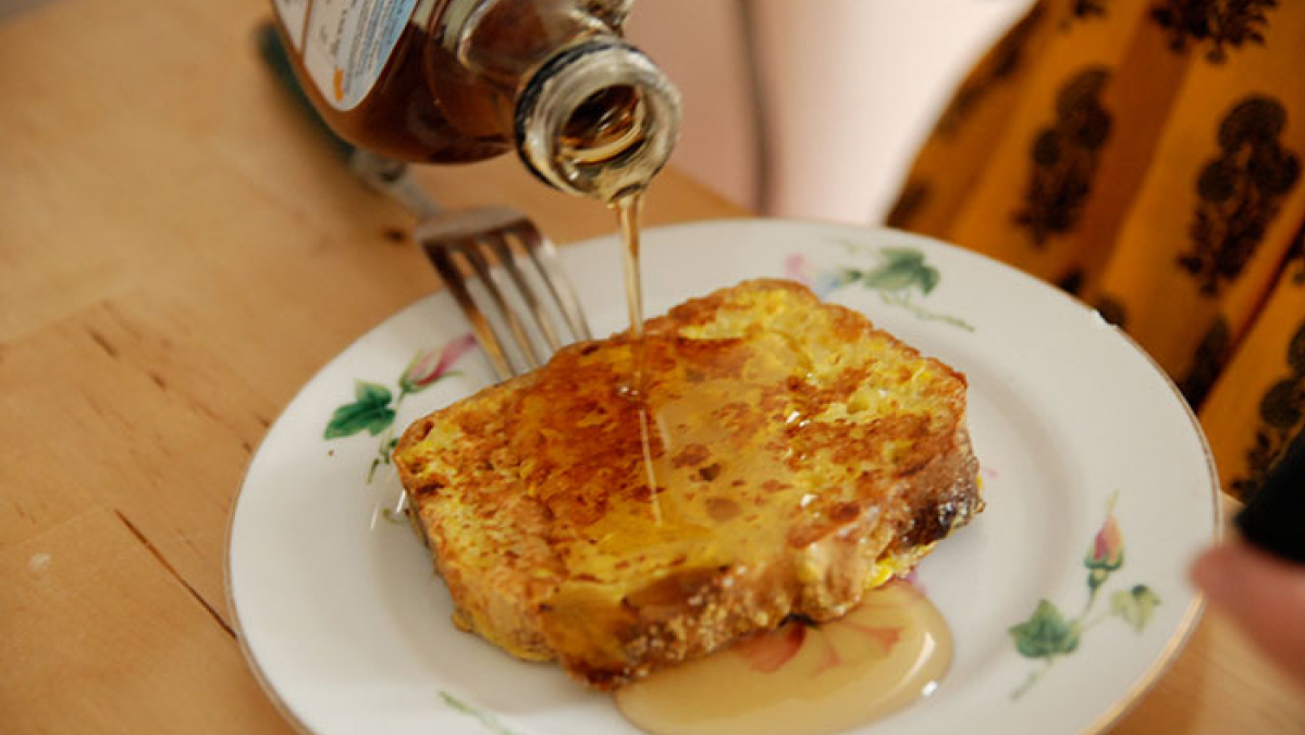 Pouring syrup over a slice of French toast on a floral plate representing national stereotypes about breakfast habits