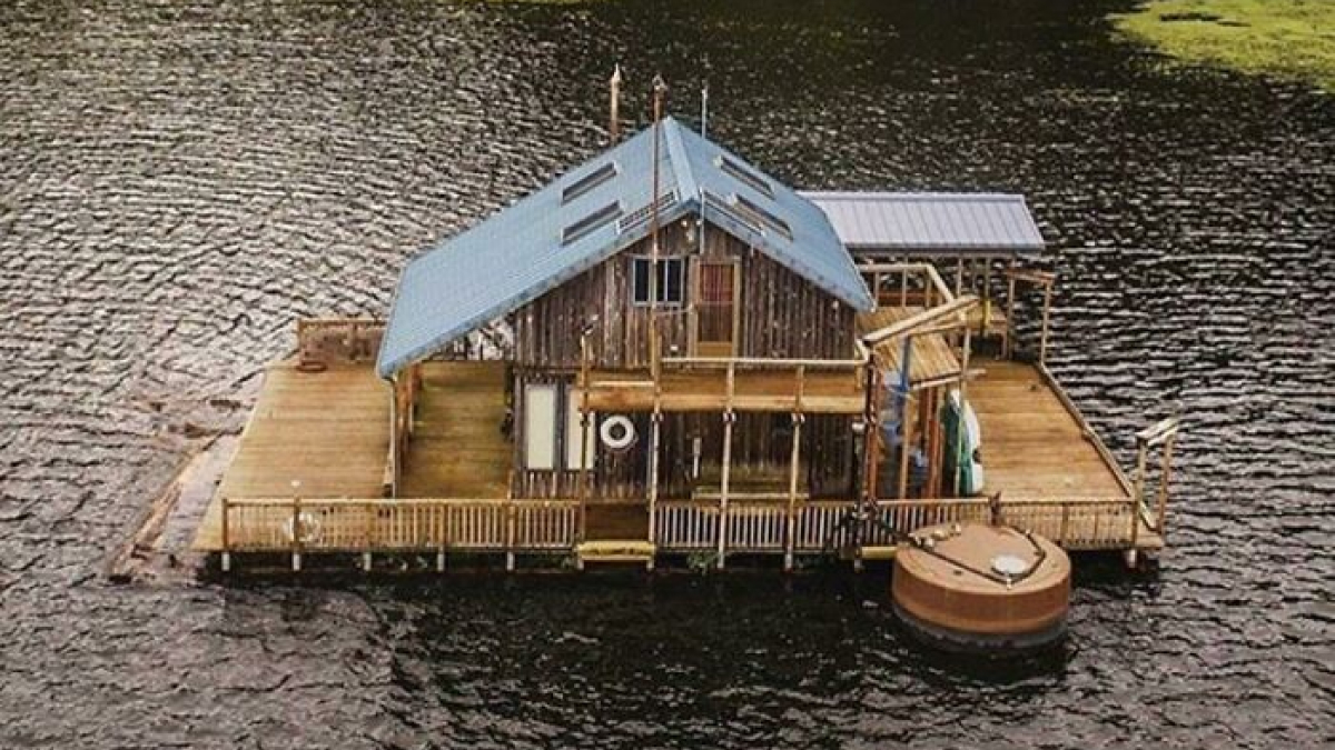 Unique tiny home floating on water with wooden decks and blue roof, showcasing off the grid living and eco-friendly design.