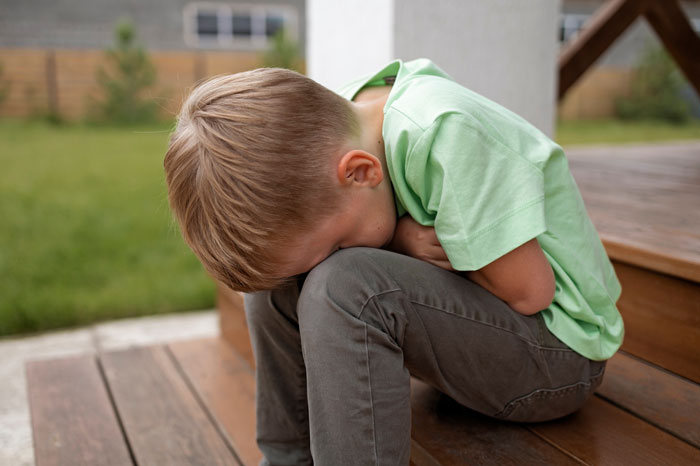 Young boy sitting on wooden steps with head down, representing neighbor’s kid turning fence into fast food window for dogs.