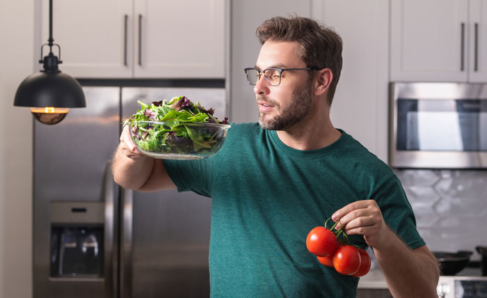 Man in glasses holding a bowl of salad and tomatoes in kitchen, reflecting on secret scale for measuring number twos. Man in glasses holding a bowl of salad and tomatoes in kitchen, reflecting on secret scale for measuring number twos.