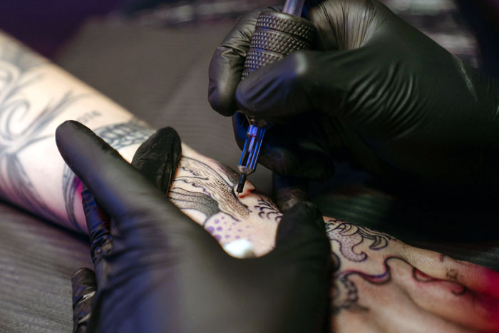 Tattoo artist working on detailed arm tattoo as man demands his girlfriend remove tattoo honoring late boyfriend. Tattoo artist working on detailed arm tattoo as man demands his girlfriend remove tattoo honoring late boyfriend.